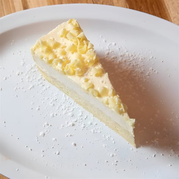 A slice of white chocolate cheesecake with white chocolate curls and powdered sugar on a white plate.