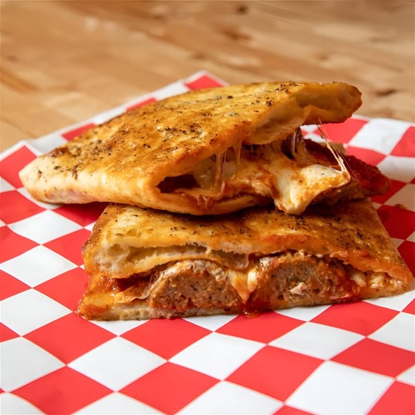 Two golden-brown calzones, one cut open to reveal melted cheese and meatballs, on red and white checkered paper.