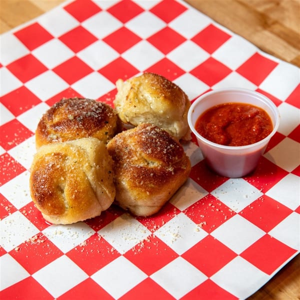 Four golden-brown garlic knots with parmesan cheese, served with a side of marinara sauce on checkered paper.