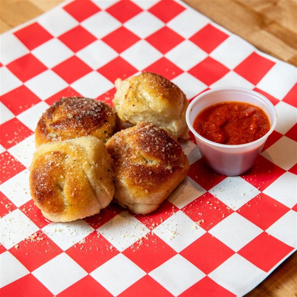 Four golden-brown garlic knots with parmesan cheese, served with a side of marinara sauce on checkered paper.