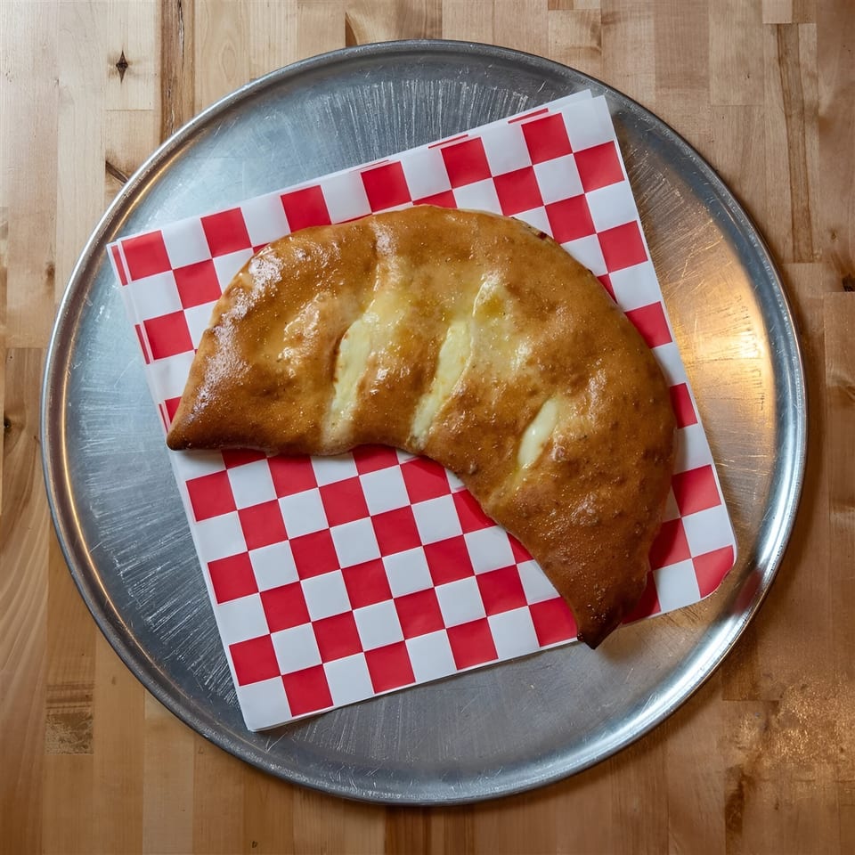Golden-brown calzone with melted cheese on red and white checkered paper, on a metal pizza tray.